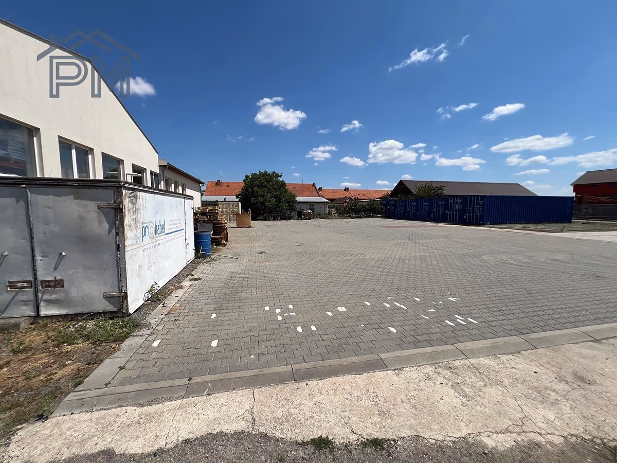 Dlážděná plocha směrem k řadě kontejnerů, vlevo skladový kontejner s paletami