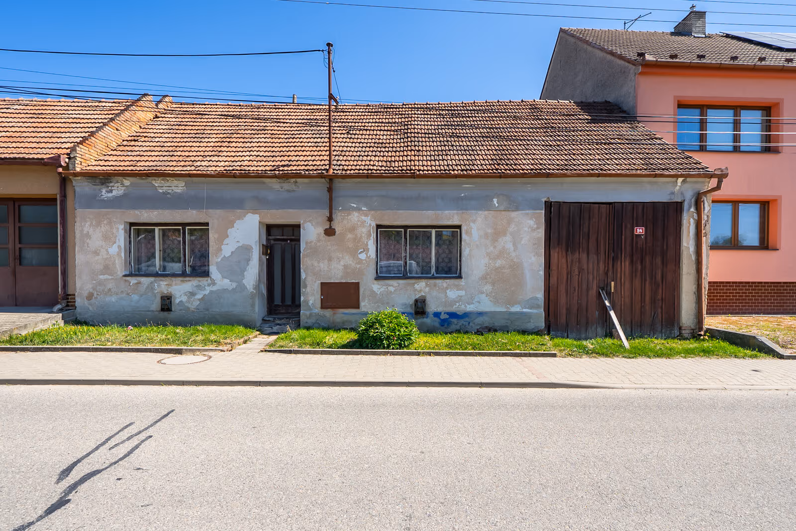 Uliční průčelí staršího přízemního řadového domu k demolici s oprýskanou omítkou, dřevěnými vraty a číslem 94, vpravo sousední zrekonstruovaný patrový dům