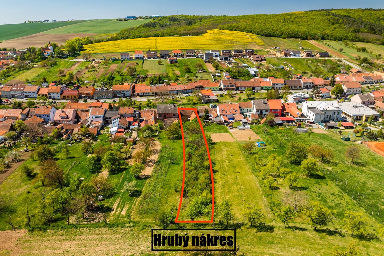 Letecký snímek obce s červeně vyznačeným orientačním obrysem pozemku (hrubý nákres) mezi zástavbou a zahradami, v pozadí řepkové pole a zalesněný kopec
