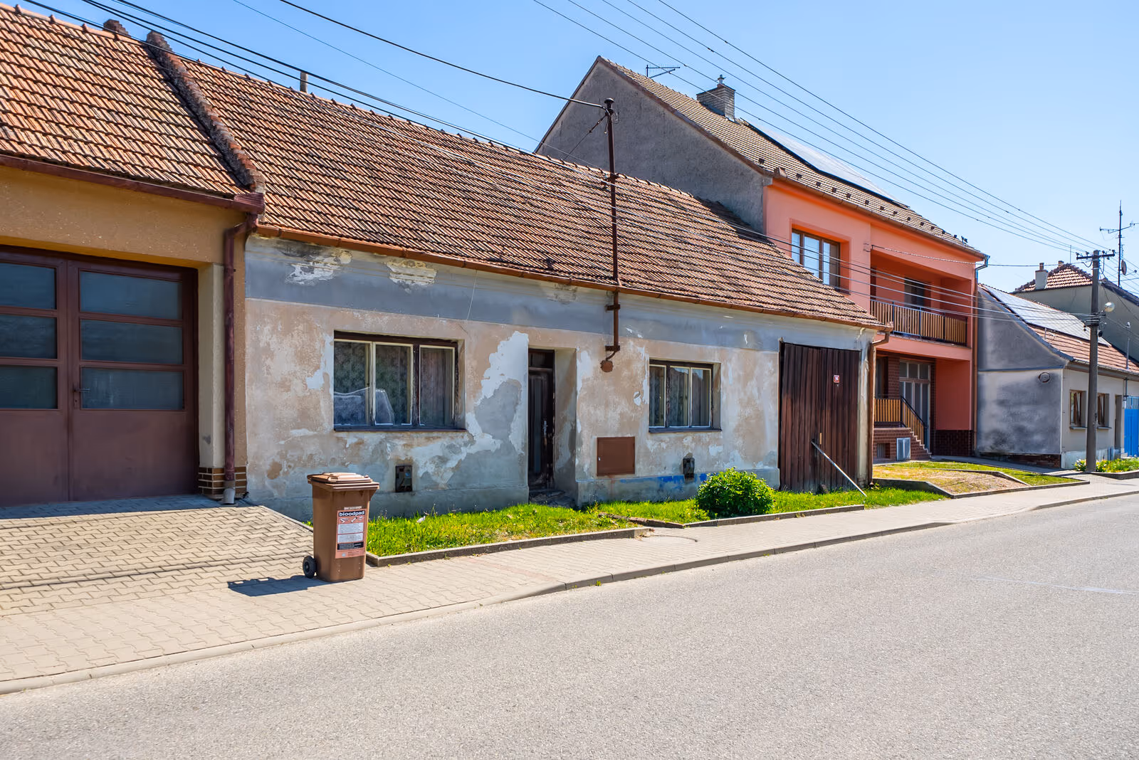 Pohled na starší dům z boku — vlevo sousední garážová vrata a hnědá popelnice na bioodpad, vpravo patrový dům, obecní silnice a chodník