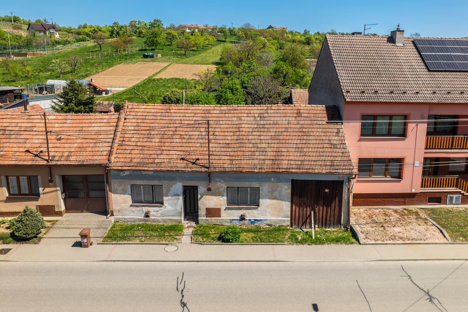 Letecký pohled na dům z druhé strany silnice — celé uliční průčelí, za domem hloubka pozemku stoupající k zahradám a návrší nad obcí