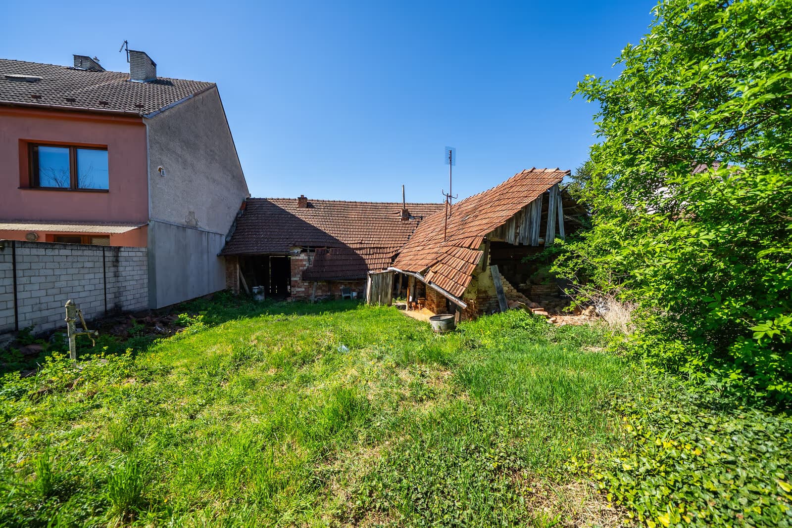 Dvůr a zahrada za domem se zarostlou travnatou plochou, vpravo křoviny, vzadu zchátralé hospodářské budovy k demolici, vlevo litinová ruční pumpa u zdi
