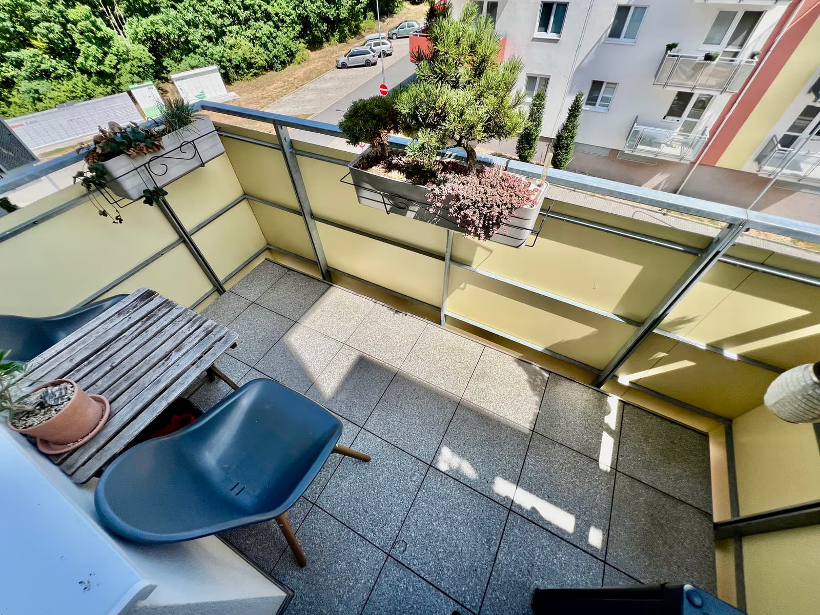 Balkon s dlažbou, dřevěným stolkem, židlí a truhlíky, s výhledem do ulice a zeleně