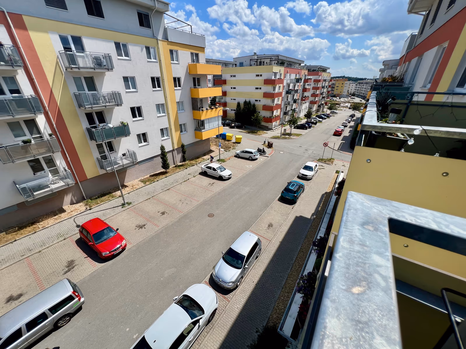 Pohled z balkonu podél ulice s řadou zaparkovaných aut a bytovou zástavbou