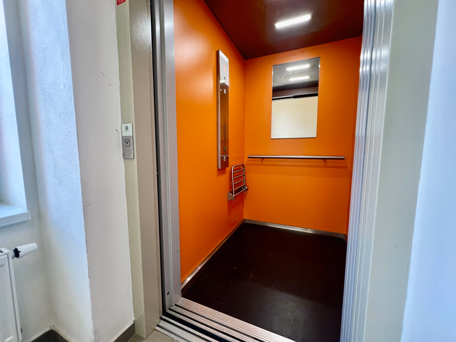Kabina výtahu s oranžovými stěnami, zrcadlem a madlem